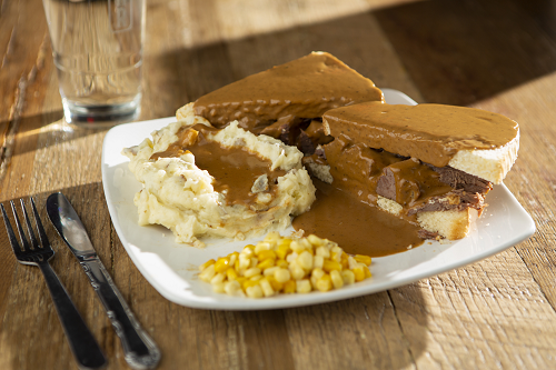 Sliced roast beef sandwich between white bread topped with homemade brown gravy served with mashed potatoes and veggies.